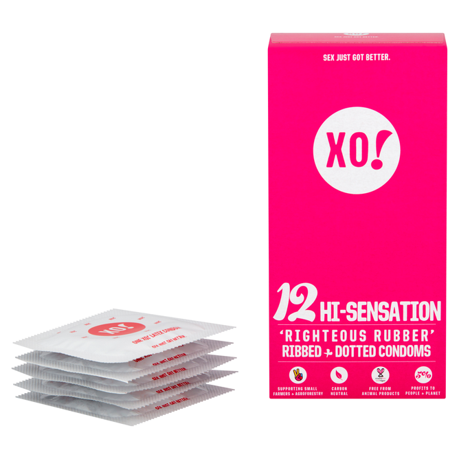 Beside a bright pink XO! box from Here We Flo, displaying 12 Hi-Sensation Righteous Rubber Ribbed + Dotted Condoms, white condom wrappers are stacked. The box highlights vegan condoms made with sustainably sourced rubber to support sexual happiness.