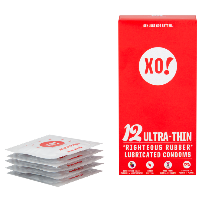 A stack of individual condom packets sits beside an upright red box labeled XO! Ultra-Thin Righteous Rubber Condoms by Here We Flo, crafted from sustainably sourced rubber, promoting sexual health, carbon neutrality, and offering a nearly naked sensation without using animal products.