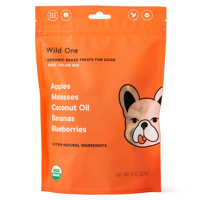 The bright orange Wild One bag features a cartoon dog and contains Organic Fruit Salad Mix Baked Dog Treats. These USDA organic treats include apples, molasses, coconut oil, bananas, blueberries, and more in an 8-ounce package with the seal on the bottom left.