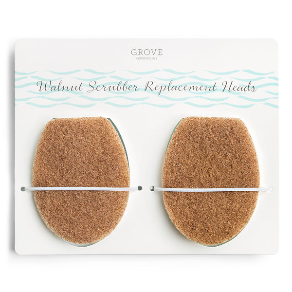 A pack of two oval-shaped, non-abrasive Grove Co. Walnut Scrubber Brush Replacement Heads on a card with a blue wave design and Walnut Scrubber Replacement Heads text, each secured by a white elastic band.