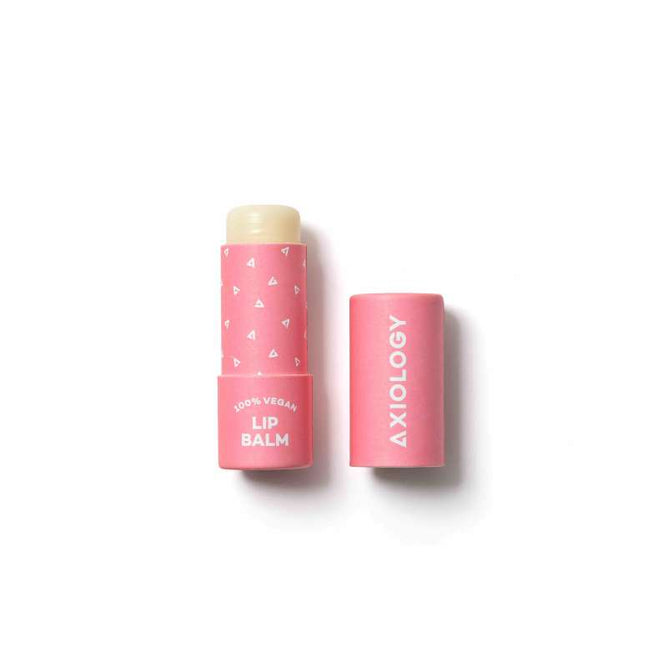 A pink tube of Axiology Lip Balm with naturally-derived ingredients like cocoa butter is uncapped, displaying the balm. The packaging includes white triangles and 100% VEGAN LIP BALM, while the cap shows AXIOLOGY.