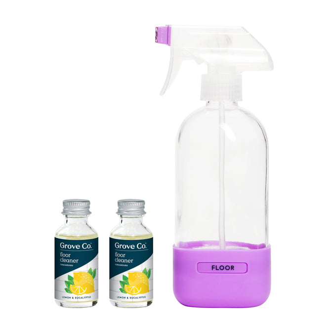 A clear spray bottle with a purple base labeled FLOOR is next to two small bottles marked Grove Co. featuring lemon and eucalyptus images. This Floor Cleaner Concentrate Starter Pack offers a reusable cleaning spray with plant-based ingredients for an eco-friendly clean.