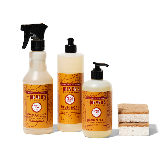 The Grove Collaboratives Happy Harvest Essentials Set features three Mrs. Meyers Clean Day apple cider-scented products—a multi-surface spray, dish soap, and hand soap—accompanied by two stacked beige sponges on the right.