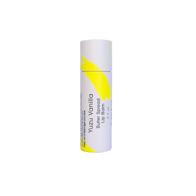 The white tube features an eco-friendly yellow abstract design and the text Butter Spread Lip Balm by butter & me - Yuzu Vanilla Blue Spruce, 0.3 oz. Perfect for soothing chapped lips, it stands upright on a plain white background.
