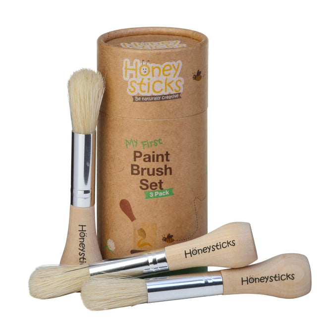 The Honeysticks My First Paint Brush Set features a 3-pack of natural bristle brushes with ergonomic wooden handles, displayed in a cylindrical cardboard container adorned with bee and honeycomb illustrations—ideal for little artists first strokes.