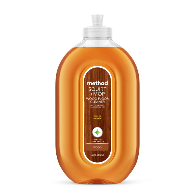 The Squirt + Mop Wood Floor Cleaner by method, with an almond scent, is ideal for hardwood floors. It comes in a clear bottle displaying amber liquid, featuring a nozzle cap and a label with product details and wood texture design to highlight its effectiveness as a wood floor cleaner.