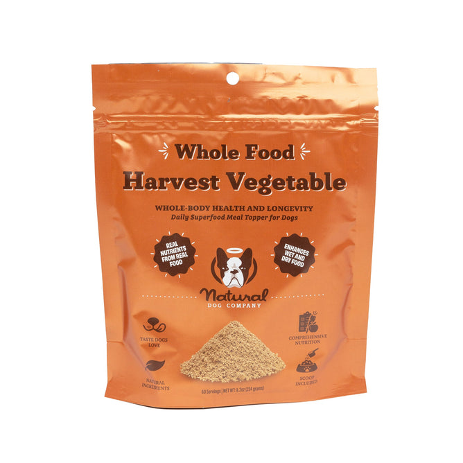 The orange packaging of Natural Dog Companys Harvest Vegetable Superfood Granules showcases its meal topper benefits, with text highlighting nutritional advantages and icons for taste, quality confidence, and digestive health. It also features an image of a dog and playful paw prints.