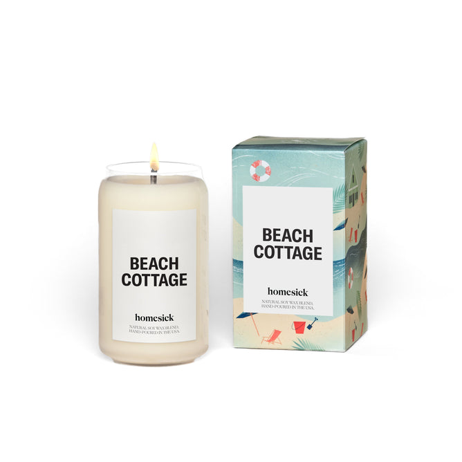 A lit Beach Cottage soy wax candle by Homesick rests next to a decorative box with beach illustrations like umbrellas and chairs. The white candle has a minimalistic label with black text on a plain backdrop.