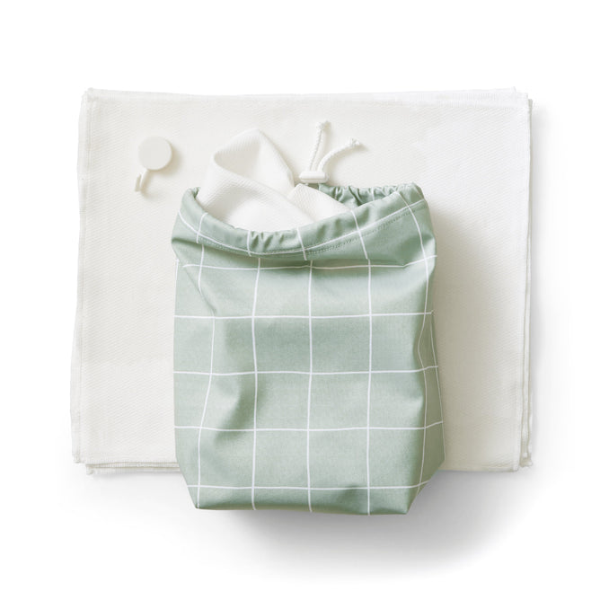 A green organic cotton drawstring bag with a white grid pattern rests on a white cloth, accompanied by a small round white object. The open bag hints at hidden treasures from Esembly Babys Reusable Paper Towels Set.