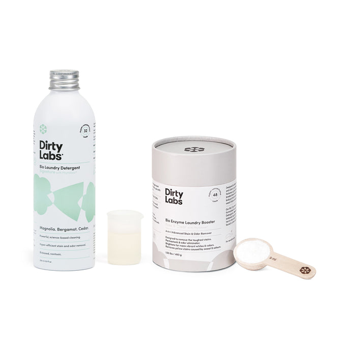 A bottle of Dirty Labs Bio Laundry Detergent with Phytolase® sits beside a cylindrical Bio Enzyme Laundry Booster, featuring an open lid and wooden scoop. This enzyme-powered duo offers a minimalist, eco-friendly design ideal for sustainable cleaning.