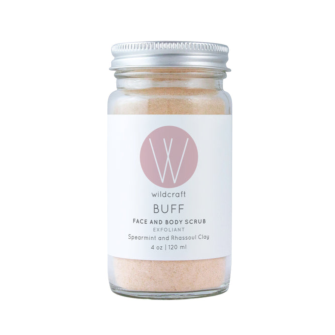A 4 oz (120 ml) glass jar of Wildcraft Buff Face and Body Scrub with a metal lid features a white label with a pink W logo, describing it as an exfoliant containing spearmint and rhassoul clay.