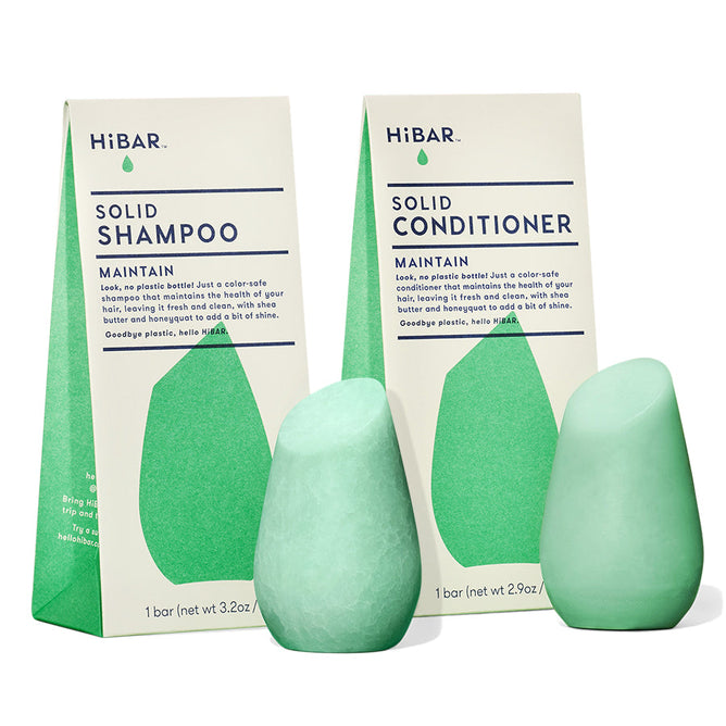 Two upright, teardrop-shaped HiBAR Maintain Solid Shampoo & Conditioner bars with shea butter, in light green, stand before their triangular packaging. Their minimal design highlights a commitment to a plastic-free, eco-friendly message while remaining color-safe.