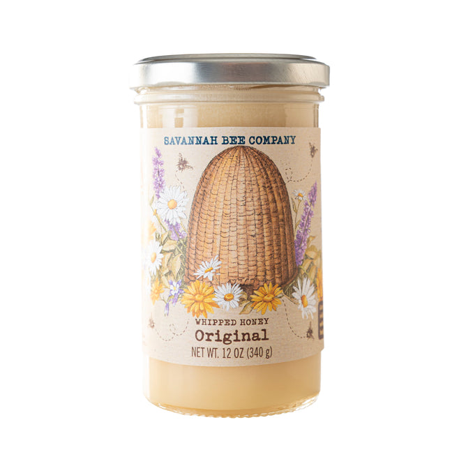 Savannah Bee Companys Whipped Honey Original comes in a 12 oz (340 g) clear glass jar, ideal for baked goods. The label displays a skep with illustrated flowers and bees, and the creamy honey inside is light in color.