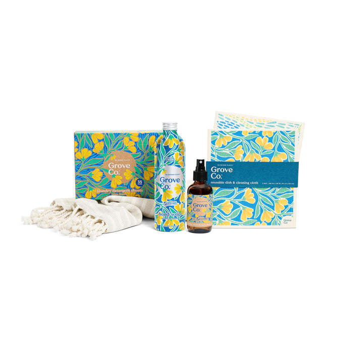 The Coastal Clean Set from Grove Collaboratives Spring Collection features limited edition Pacific Primrose fragrances. The set includes a bottle, spray, and cleansing cloths with floral packaging, artfully placed next to a folded beige towel with fringed edges.