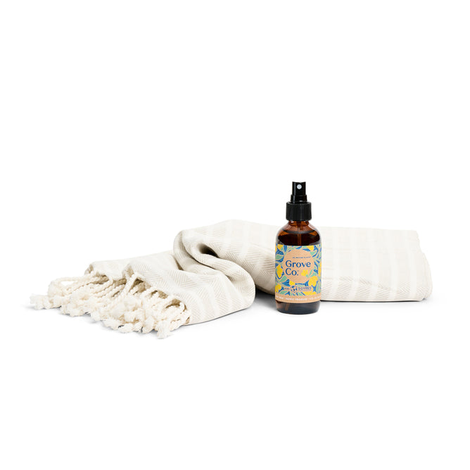A Grove Collaborative Bathroom Refresh Set - Spring Collection bottle with a spray nozzle is on a white background beside a folded cream towel with tassels. The bottle boasts a colorful, patterned label.