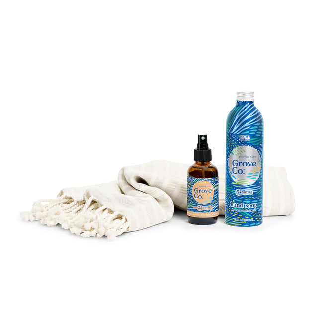A striped woven towel with fringed edges is folded next to two Grove Collaborative Hostess Gift Set products from the Spring Collection. One is a spray bottle and the other a larger bottle, both featuring blue labels with artistic wave designs and limited edition scents like Atlantic Dune Grass.