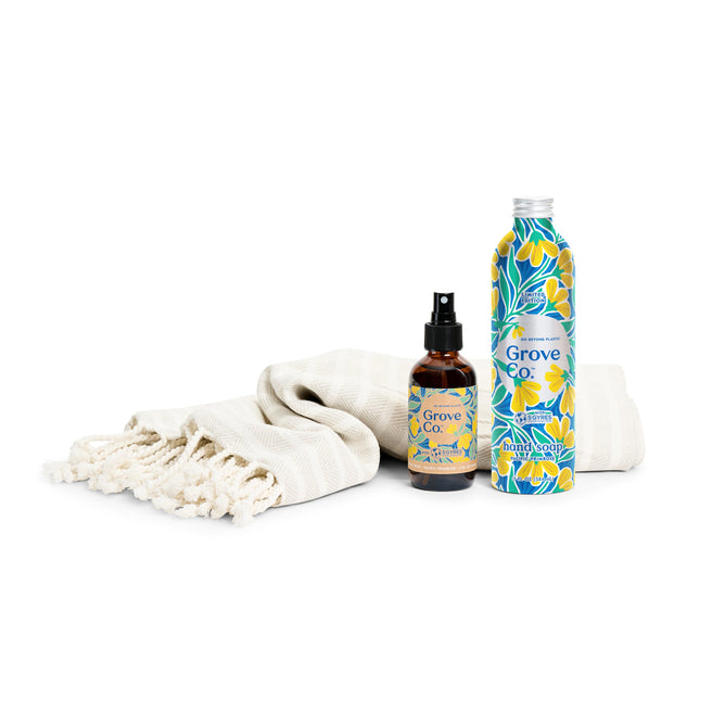 A beige fringed towel sits beside Grove Collaboratives Hostess Gift Set from the Spring Collection, featuring a spray bottle with a yellow and blue label, and a metal bottle adorned in the Pacific Primrose pattern with blue, white, and yellow floral designs.