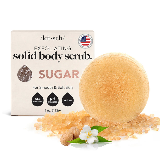 The image displays the Kitsch Sugar Exfoliating Solid Body Scrub, a round scrub labeled sugar. The box in the background notes that its all-natural, pH balanced, and vegan. A white flower and nutmeg accompany scattered scrub, giving off caramel and vanilla bean hints.