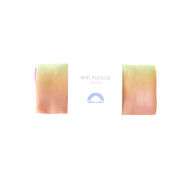 A Mini Playsilk by Sarahs Silks, made from 100% pure silk, features a pastel gradient of pink, peach, and green. It includes a label with MINI PLAY SILK ENCHANTED and a rainbow logo above SARAHS SILKS. Ideal for open-ended imaginative play.