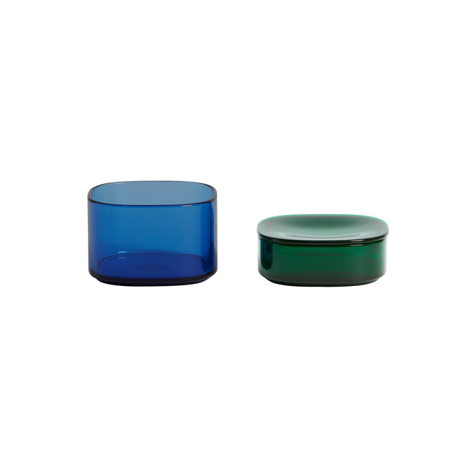 Two cylindrical Storage Gems by Open Spaces are displayed side by side. The taller left container, resembling crystalline acrylic, is blue, while the shorter right one has a rounded lid and vibrant green hue. Both feature a sleek, modern design.