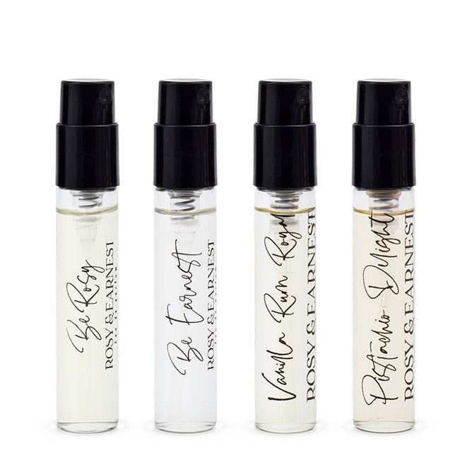 Four small spray bottles from the Rosy & Earnest Discovery Set are displayed in a row. Each has a black cap and labels with scent names: Be Rosy, Be Earnest, Vanilla Rum Rosy—featuring smooth vanilla and bold rum—and Perfumers Delight.