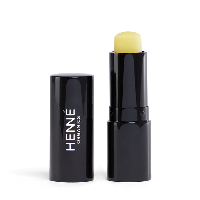 A plain white background features a black tube labeled in white with Henné Organics, a women-owned, natural skincare brand. The cap is off, revealing its pale yellow Luxury Lip Balm.