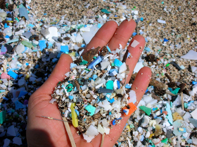 A hand holding a mix of small, colorful plastic fragments and pebbles on sandy debris spotlights the critical issue addressed by Grove Co.s Protect People and Planet from Microplastics.