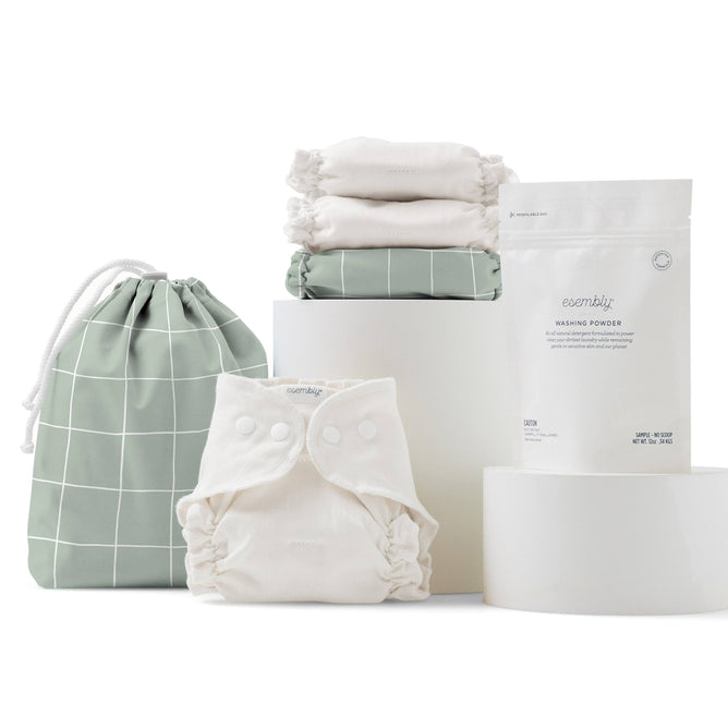 Discover the eco-friendly Esembly Baby Cloth Diaper Try-It Kit, Size 1, elegantly displayed with a green grid-patterned drawstring bag, organic cotton white cloth diapers, a white snap-button diaper, and washing powder on sleek white cylindrical platforms.