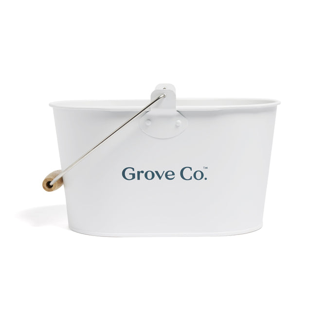 The Matte Cleaning Caddy by Grove Co. is a white, oval storage caddy with a wooden handle and metal supports. Its ideal for organizing cleaning products or art supplies, featuring Grove Co. printed in blue on its side.