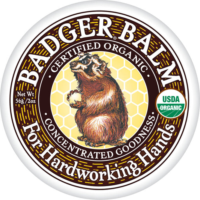 A round tin of Badger Balm for Hardworking Hands showcases a badger illustration with a honeycomb background. The labels feature Certified Organic, Concentrated Goodness, and the USDA Organic logo, emphasizing its organic ingredients. Net weight: 2 oz.
