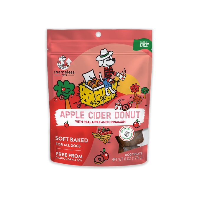 The packaging for Shameless Pets Apple Cider Donut dog treats showcases a cartoon dog in a wagon surrounded by cinnamon donuts, apples, and cinnamon sticks. These 6 oz (170g) soft-baked treats are grain, corn, and soy-free and are proudly made in the USA.