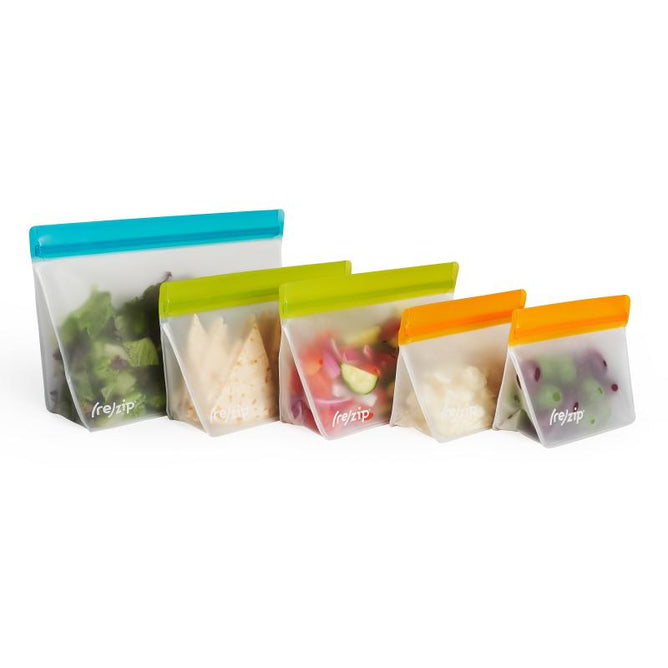 The (re)zip Stand-Up Pack n Go Kit features five freezer-safe reusable silicone food storage bags in various sizes, with colorful tops in blue, green, yellow, and orange. They are displayed on a white background containing assorted foods such as lettuce and berries.