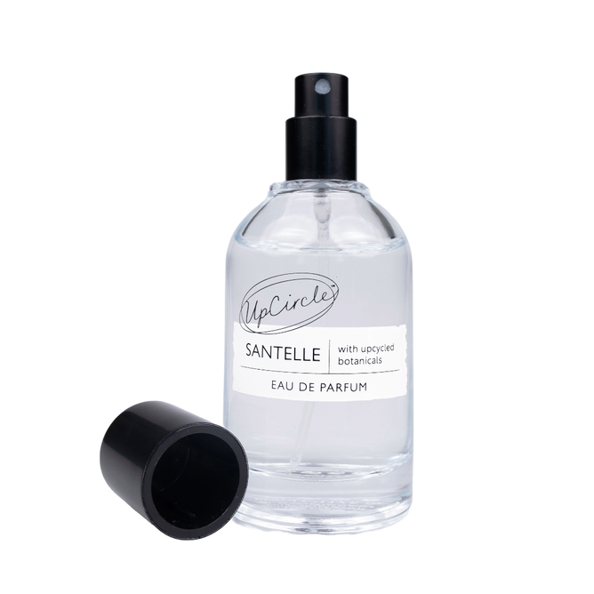 A clear glass perfume bottle labeled UpCircle Upcycled Eau de Parfum features a black spray nozzle and uncapped black lid. The design highlights with upcycled botanicals, showcasing the essence of sandalwood against a minimalist white background, enhancing its elegance.