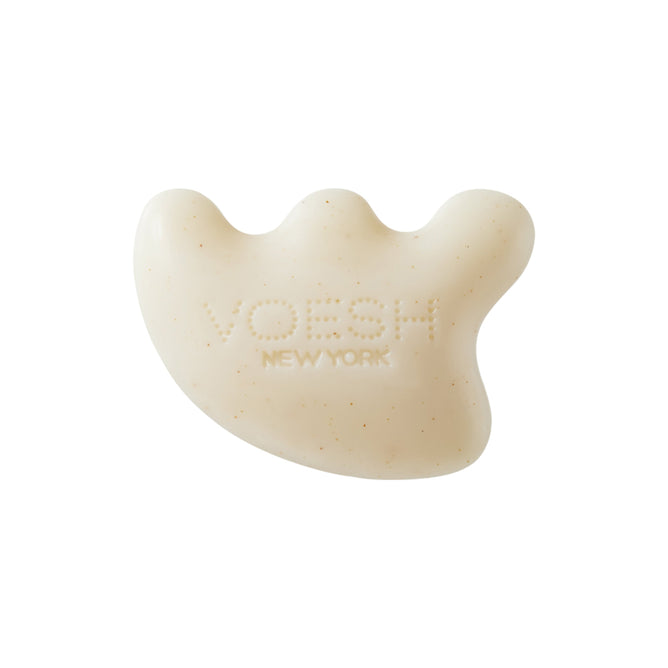 The Gua Sha Shampoo Bar by VOESH New York is a white soap with a unique shape, featuring three rounded protrusions and slight speckling. It promotes hair growth while adding an elegant touch with its smooth texture and branding.