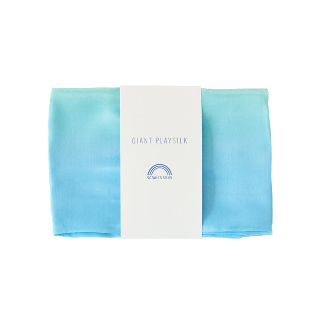 The Giant Playsilk by Sarahs Silks, ideal for open-ended play in Montessori and Waldorf homes, displays a gradient from light blue to turquoise. It is wrapped with a white paper band labeled Giant Play Silk featuring a small rainbow graphic.