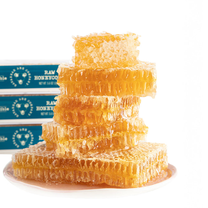 A stack of raw honeycomb glistens with golden Hungarian honey on a white plate, while blue and white packages labeled Raw Honeycomb by Savannah Bee Company provide a beautiful backdrop, showcasing honeys natural beauty.