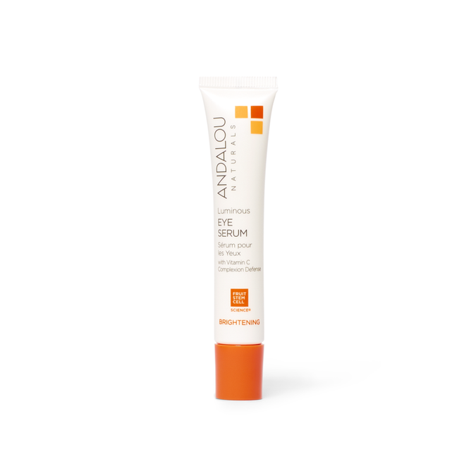 The white tube of Andalou Naturals Brightening Luminous Eye Serum, with an orange cap, highlights its brightening power using Vitamin C and Fruit Stem Cell Science, featuring an orange square logo at the top that signifies its innovative formula.