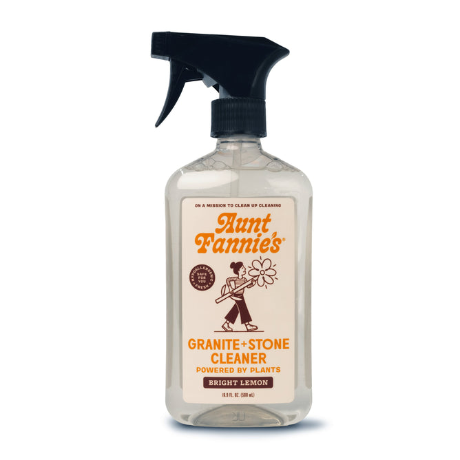 The Aunt Fannies Granite + Stone Cleaner comes in a clear spray bottle labeled Bright Lemon and Powered by Plants, featuring natural surfactants. The white label with an illustration of a person holding a flower is complemented by a black cap, and essential oils enhance its cleaning power.