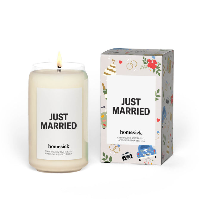 A lit Homesick Just Married candle, crafted from natural soy wax in a glass jar, features the text JUST MARRIED. It sits next to a box decorated with cakes, rings, flowers, and cameras, capturing the essence of ocean air for newlyweds.