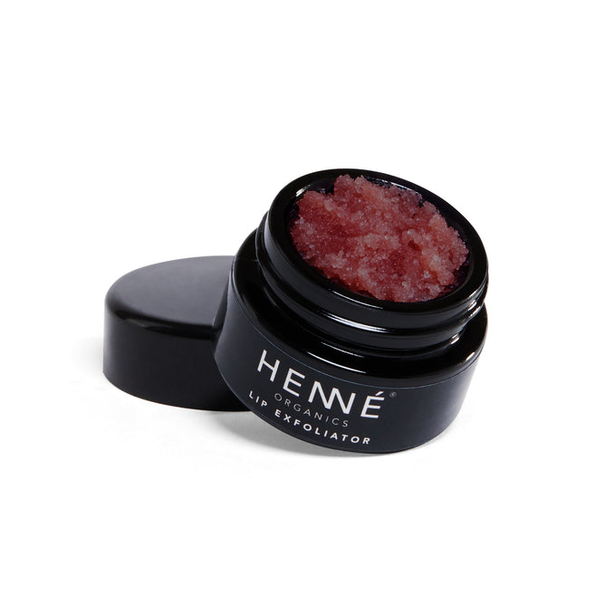 An open Henné Organics Lip Exfoliator jar reveals its textured, red formula with sugar crystals. The black jar and lid elegantly rest on a white surface, accentuating the products luxury.