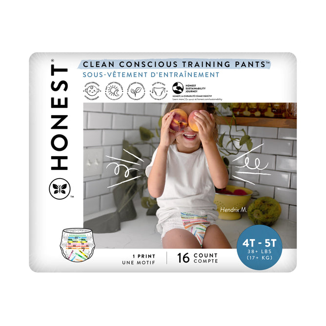 A child playfully holds orange halves over their eyes on a bathroom counter, wearing white training pants with colorful designs, ideal for potty-training. The packaging reads The Honest Company: Training Pants - 4T/5T, 38+ lbs, 16 count.