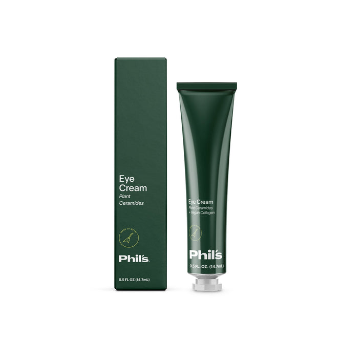A green tube of Phils Eye Cream with plant ceramides and vegan collagen is next to its matching vertically oriented box. The box has minimalist designs with white text on a dark green background, and the tube holds 0.5 FL OZ (14.7 mL).