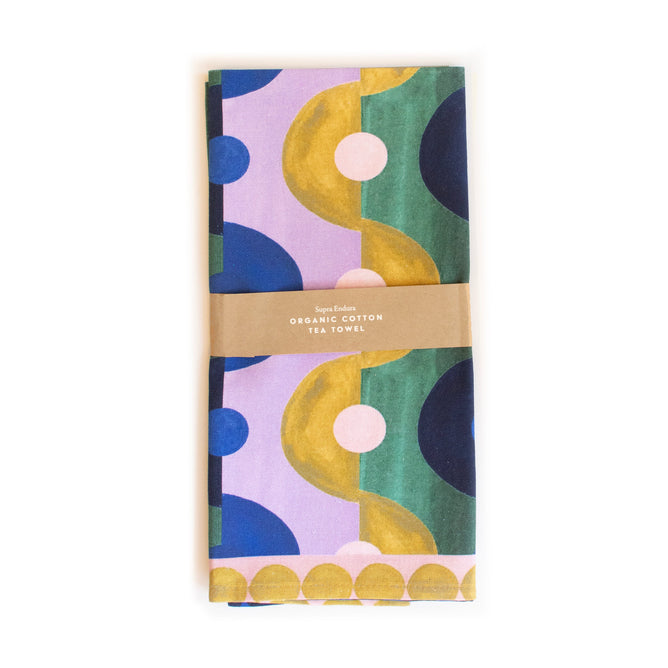 The folded Organic Cotton Tea Towel - Limited Edition from Supra Endura showcases a vibrant geometric pattern in green, blue, and gold on a lavender background. Wrapped in a brown paper band, it highlights its sustainable GOTS-certified organic cotton material.