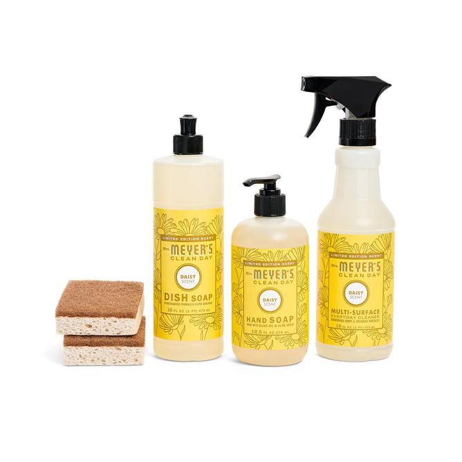 The Blooming Buds Scrubbing Set by Mrs. Meyers includes daisy-scented dish soap, hand soap, and multi-surface cleaner, along with two walnut scrubber sponges. The packaging is vibrant in yellow with lovely floral designs.