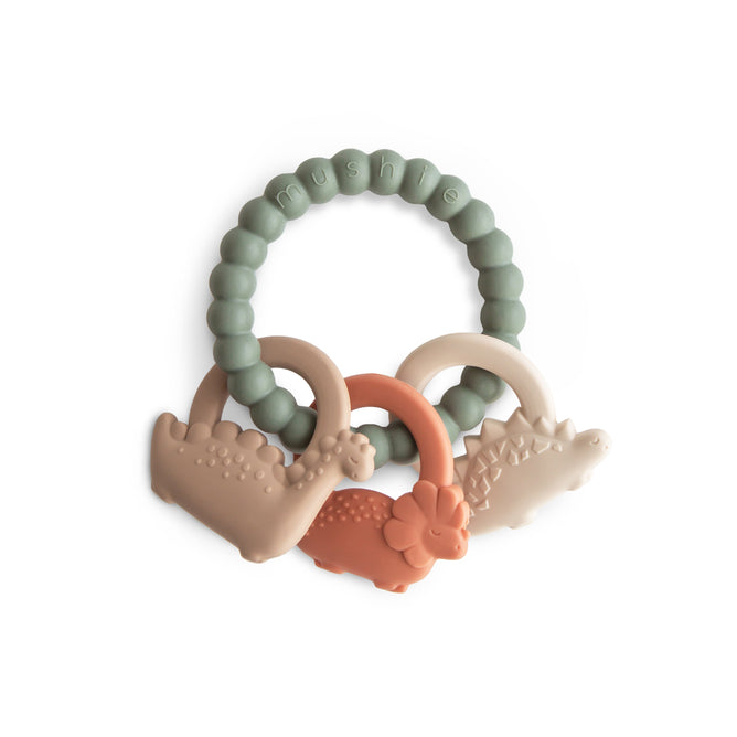 The mushie Dino Teething Ring is made of BPA-free, food-grade silicone and features a green beaded ring with three dinosaur-shaped sensory beads in beige, tan, and orange. mushie is embossed on the white background.