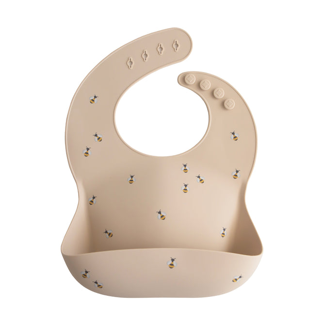 The mushie Silicone Baby Bib – Adjustable features a beige design with a small bee pattern, food-catching pocket, and adjustable snap neck closures. Made from FDA-tested food-grade silicone, it ensures safe and easy mealtimes for your baby.