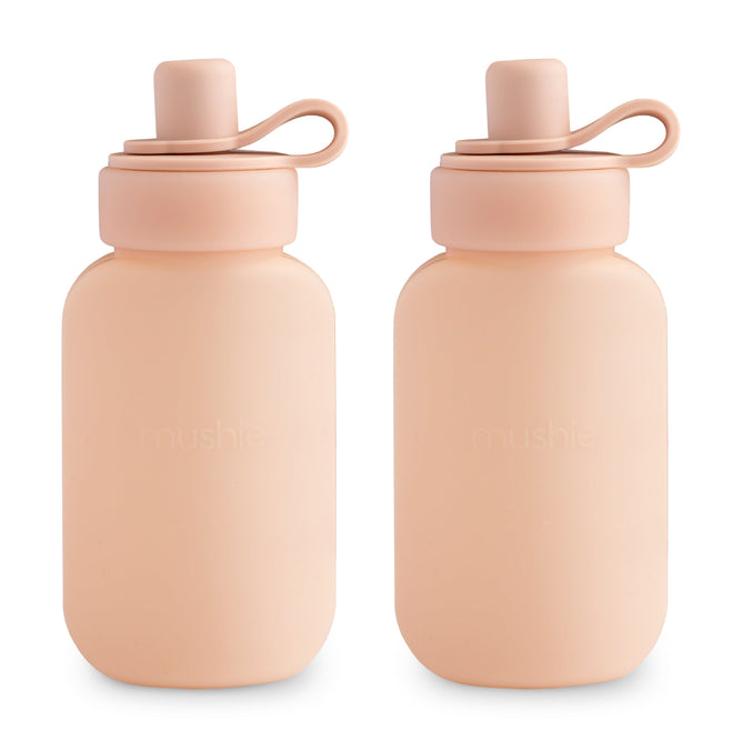 Two light peach-colored mushie Silicone Puree Pouches, reusable and BPA-free, with matching lids and looped handles, stand side by side on a white background.