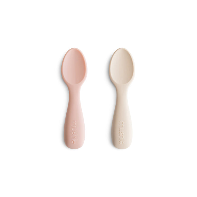 Two mushie Silicone Toddler Starter Spoons in light pink and cream, made from BPA-free, food-grade silicone, lie side by side with mushie embossed on each handle—safe and perfect for encouraging independent feeding.