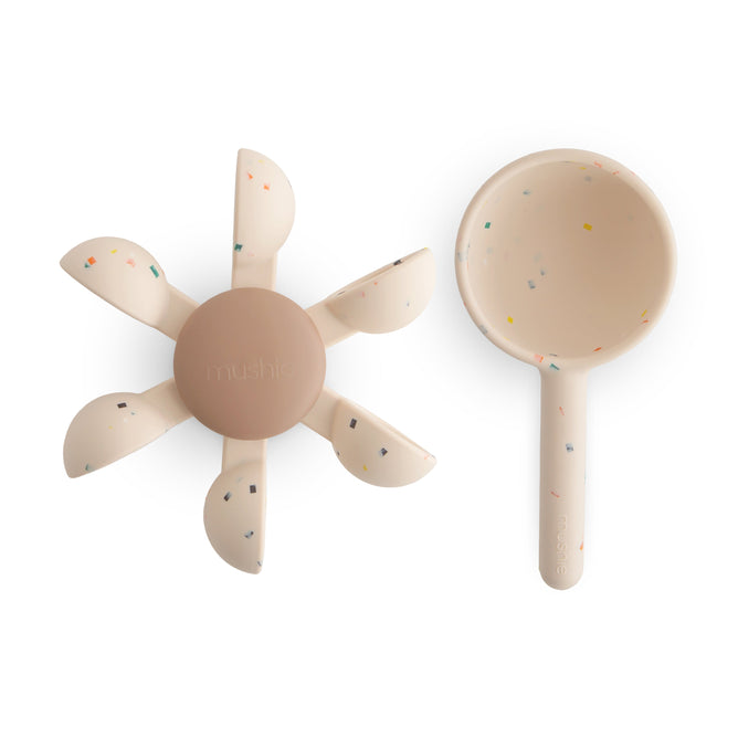 A mushie Water Wheel Bath Toy—an interactive, BPA-free bath toy in beige with colorful confetti-like specks—is shown beside a matching scoop.