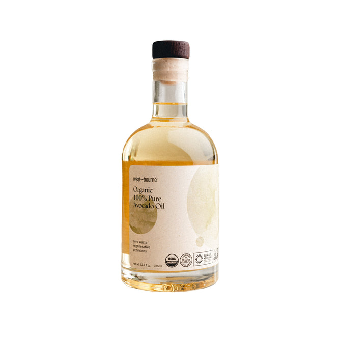 A clear glass bottle of west~bourne Organic 100% Pure Avocado Oil with a cork stopper has organic certification logos. Ideal as a finishing oil for a sustainable kitchen.